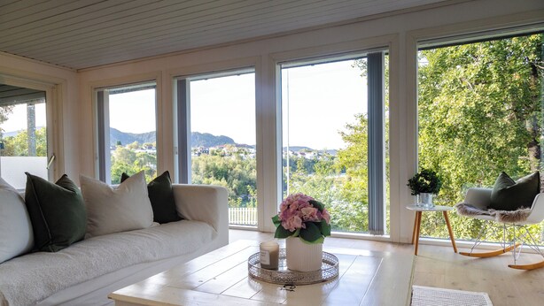 A room with a white sofa, a coffee table with a candle and flowers, and a view of greenery and hills through large windows.