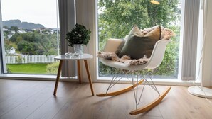 A modern room with a rocking chair, a small table with a vase, and a view of a green landscape through large windows.