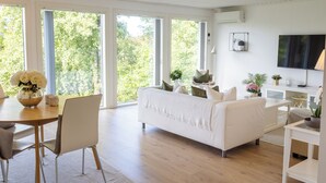 A modern living room with a white sofa, a dining table with chairs, and a TV on the wall.