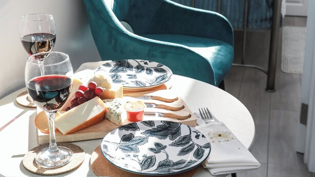 Un tavolo da pranzo con un piatto di formaggio, uva e un bicchiere di vino, una poltrona verde acqua e una struttura letto in metallo sullo sfondo.