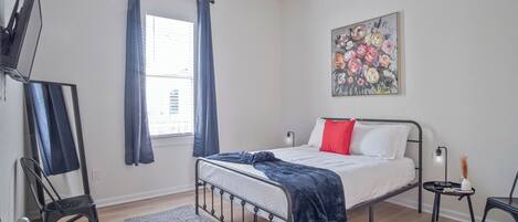 Un dormitorio con cama, un cuadro, una ventana con cortinas, una silla, una mesita y un televisor sobre un mueble.