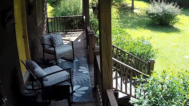 Une terrasse extérieure couverte, dotée de deux chaises et d’une table, entourée de verdure et d’une rambarde en bois.