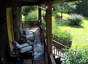 A covered outdoor deck with two chairs and a table, surrounded by greenery and a wooden railing.
