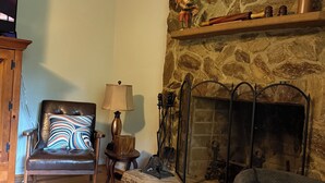 Un salon chaleureux avec une cheminée en pierre, un fauteuil en cuir agrémenté d’un coussin à rayures, un meuble en bois et un téléviseur posé sur un meuble.
