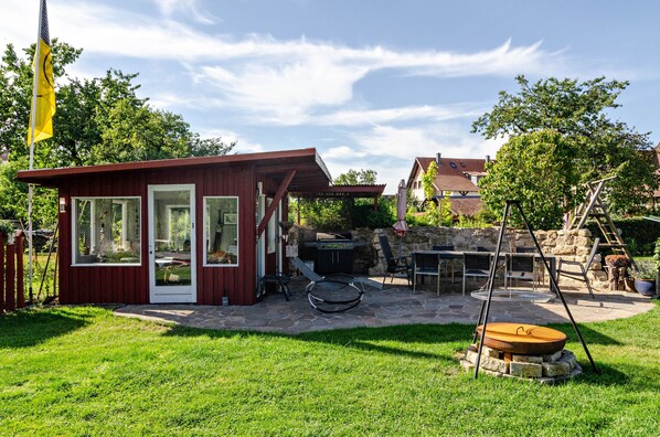 A red wooden structure with glass doors, a stone barbecue area, and a swing set.