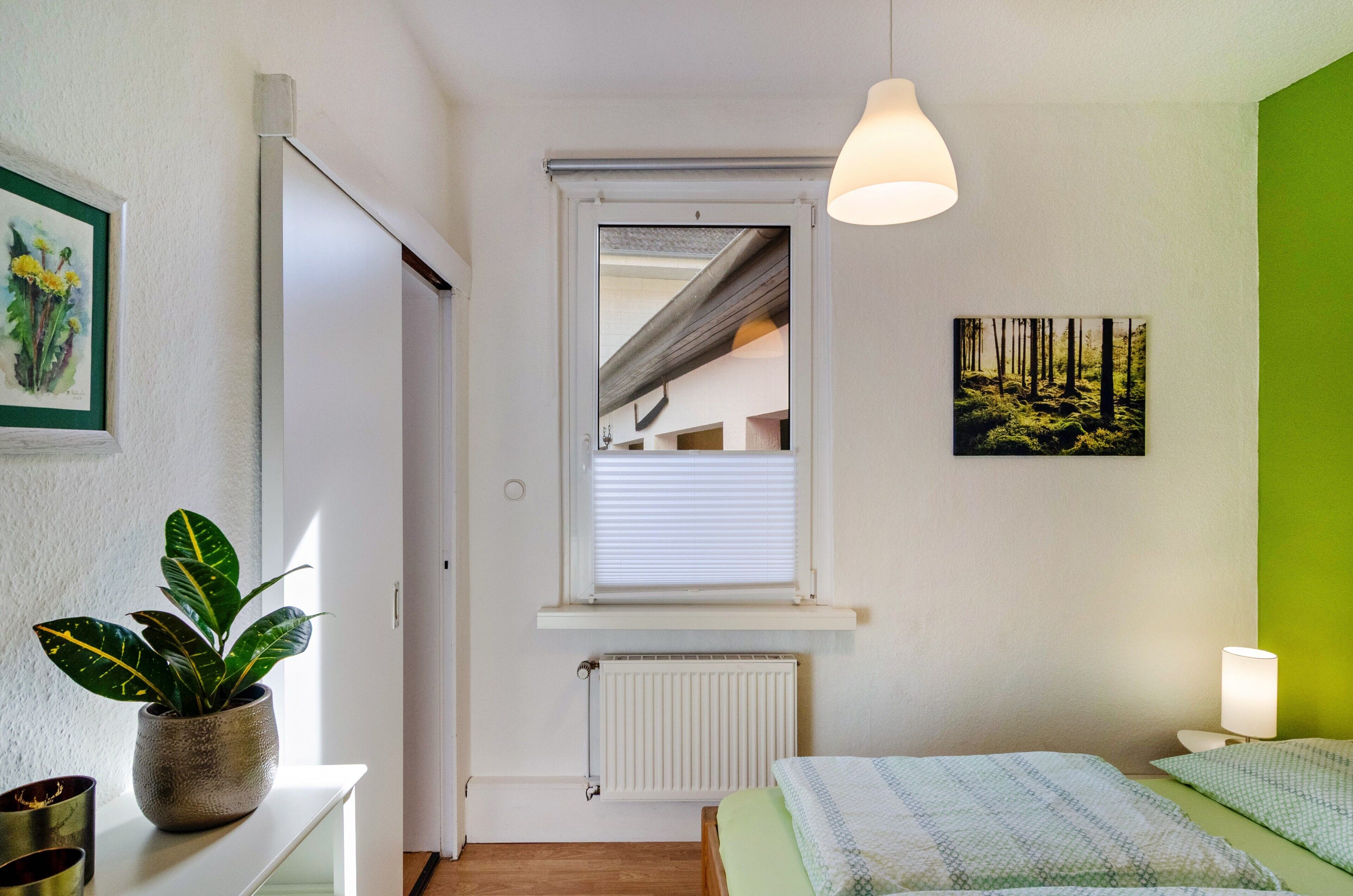 Ein Schlafzimmer mit einem Bett, einem Blumentopf, einem Fenster mit Jalousien, einer Heizung und einer grünen Wand.