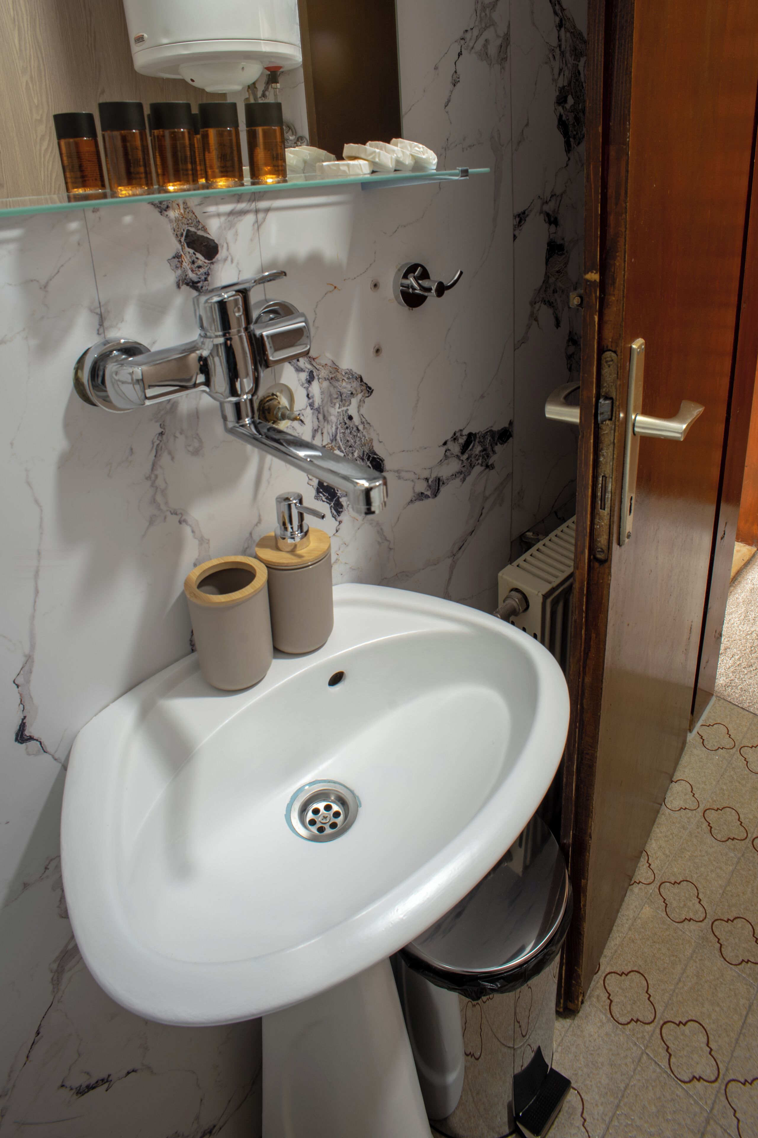 A bathroom with a white sink, a mirror, a glass shelf with toiletries, and a wooden door.