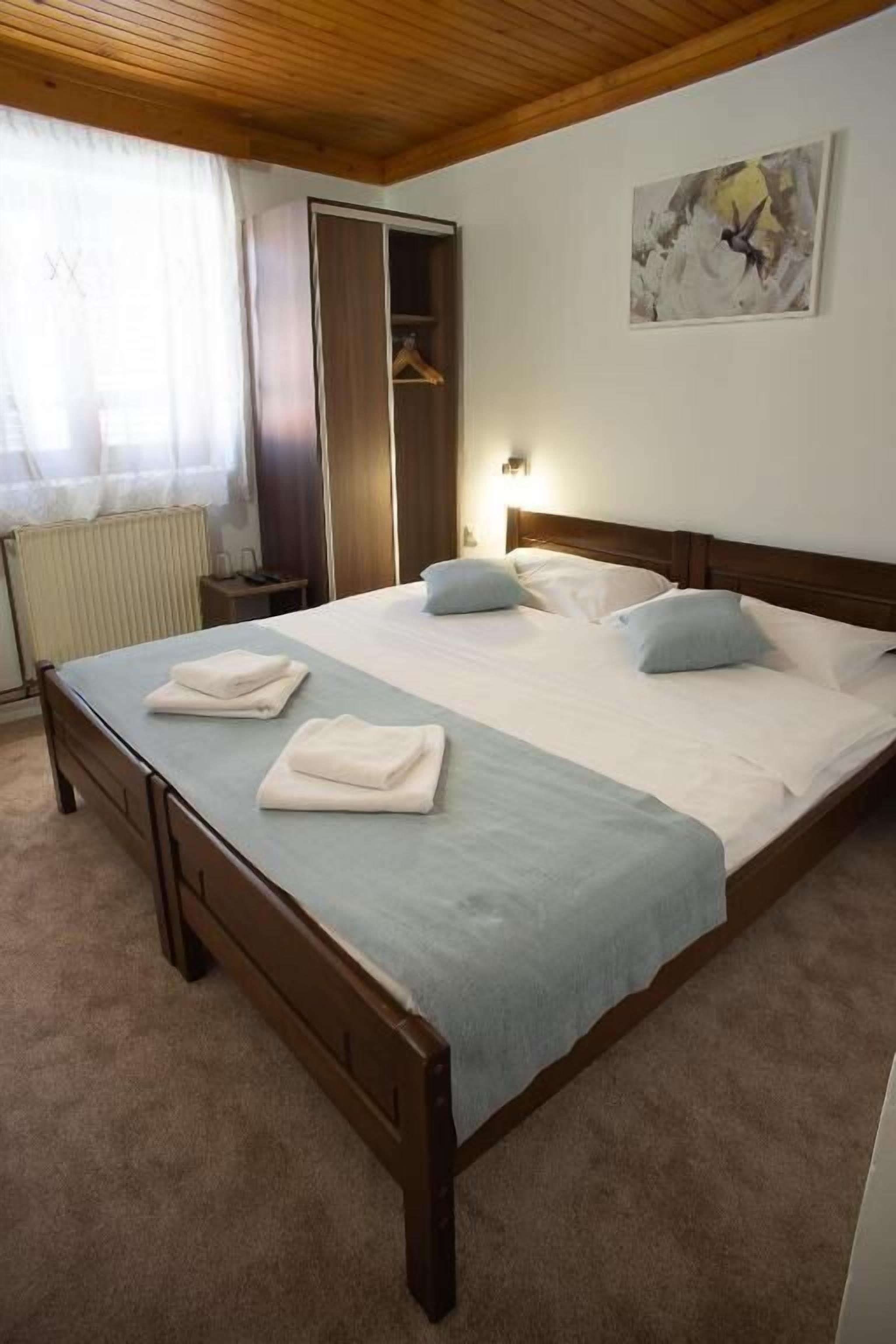 Cama bem arrumada com travesseiros, cabeceira de madeira e colcha azul claro. Há um radiador na parede e um quadro sobre a cama.