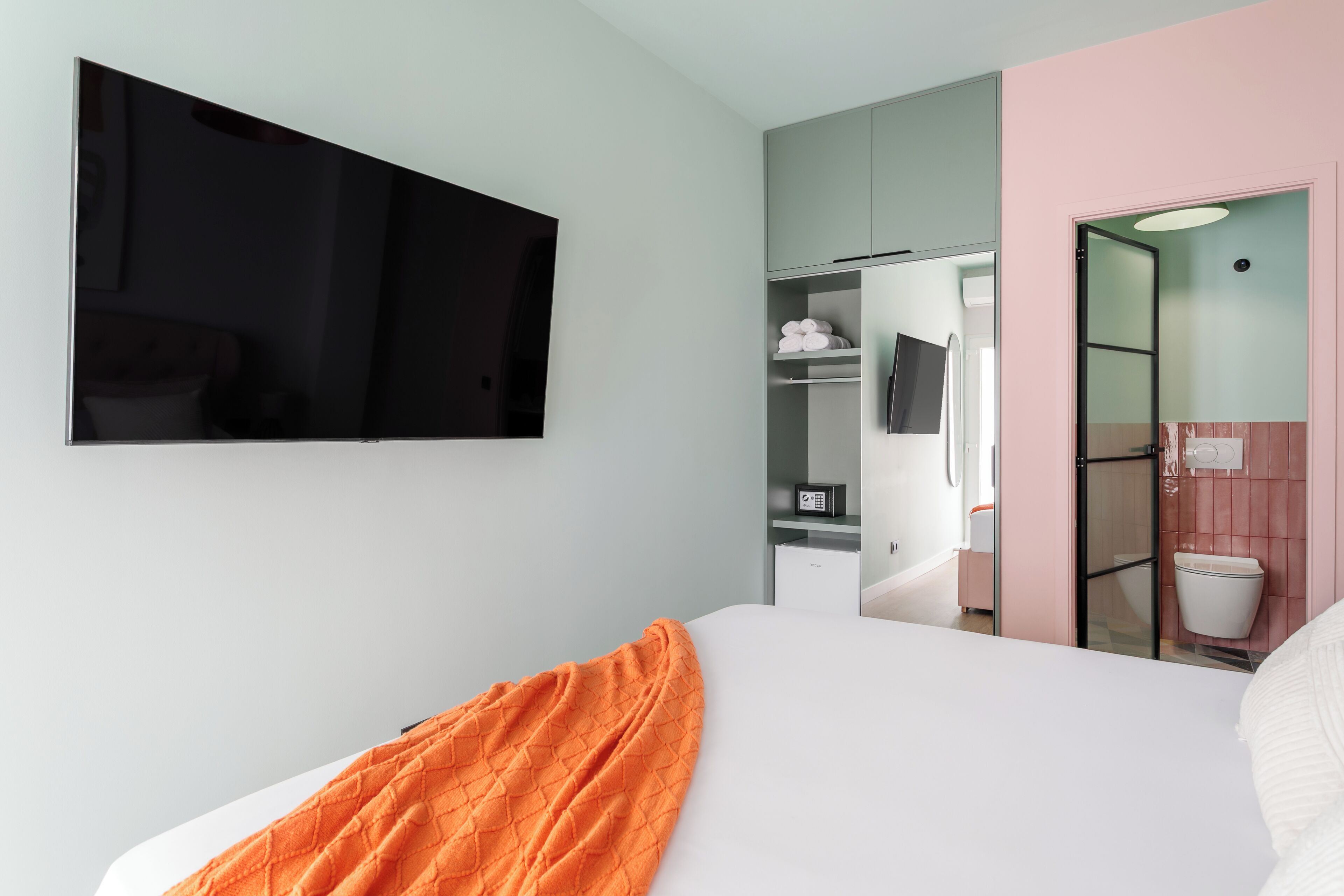 Habitación de hotel moderna con cama, televisor montado en la pared, armario y baño visible a través de una puerta abierta.
