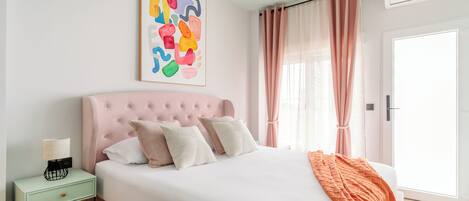 A bedroom with a pink tufted headboard, a white bedspread, and an orange throw blanket. There is a bedside table with a lamp and a framed abstract painting above it.