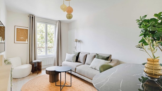 A modern living room with a sofa, a coffee table, and a side table. There is a window with curtains, a plant, and a wall-mounted lamp. - Cosy Apartment - 1bdr-4p - Ivry-sur-seine (Ivry-sur-Seine)