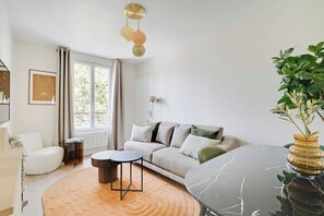 A modern living room with a sofa, a coffee table, and a side table. There is a window with curtains, a plant, and a wall-mounted lamp. - Cosy Apartment - 1bdr-4p - Ivry-sur-seine (Ivry-sur-Seine)