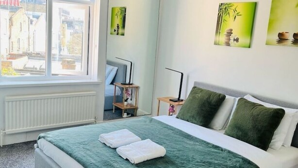 A neatly made bed with a green blanket and pillows, a bedside table with a lamp, and artwork on the wall.