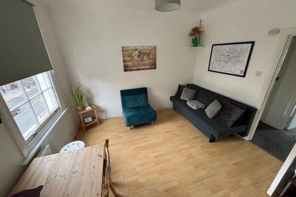 Un espacio habitable pequeño con una mesa de comedor de madera, un sillón verde azulado, un sofá oscuro y una mesita auxiliar con una planta.