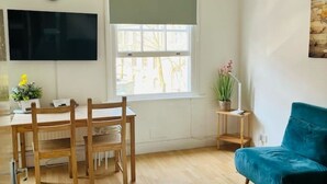 A room with a wooden dining table, chairs, a blue sofa, a TV mounted on the wall, and a window with a view of trees.
