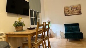 A living room with a wooden dining table, chairs, a flat-screen TV mounted on the wall, a window, and a teal sofa.