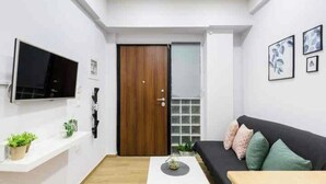 A modern living room with a flat-screen TV mounted on the wall, a wooden door, a black sofa with cushions, a white coffee table, and a small plant on a white shelf. - Charming studio in Athens 25sqm (Athina)