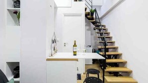 A compact kitchen with a white countertop, a wine bottle, and a small table. A wooden staircase with a metal railing leads to an upper level. - Charming studio in Athens 25sqm (Athina)
