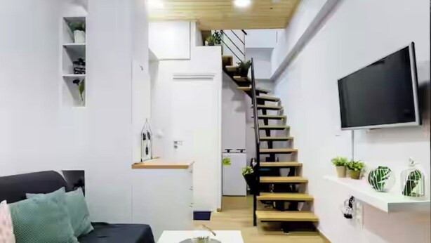 A modern living room with a black sofa, a white coffee table, and a staircase leading to a kitchen area. - Charming studio in Athens 25sqm (Athina)