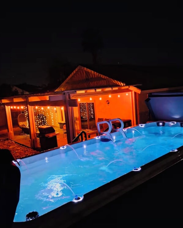 A swimming pool with lights, a covered seating area, and a building with string lights.