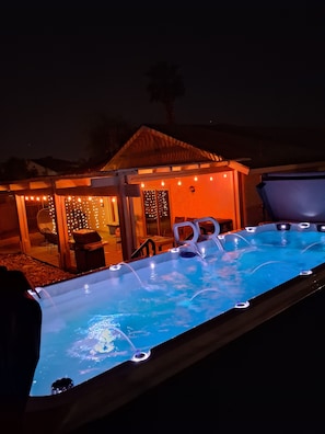A swimming pool with lights, a covered seating area, and a small building with string lights.