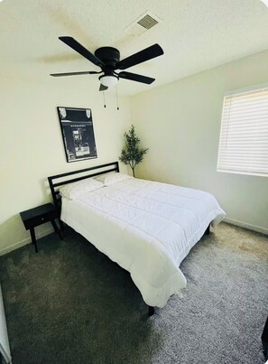 A bedroom with a bed, a ceiling fan, a small plant, a window with blinds, and a poster on the wall.