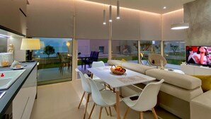 A modern living room with a dining area, a sofa, and a TV. There is a balcony with a view of the city.