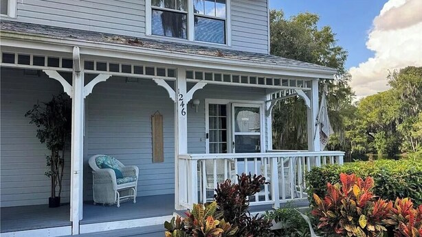 A two-story house with a covered porch, a white chair, and various plants.