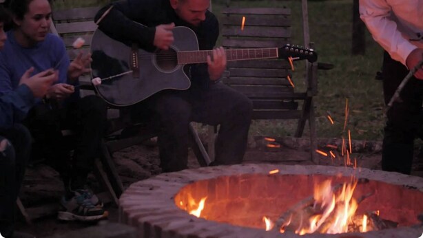 A person playing a guitar at a campfire with a group of people sitting on benches nearby. - Refugio los Volcanes (Cuevas)
