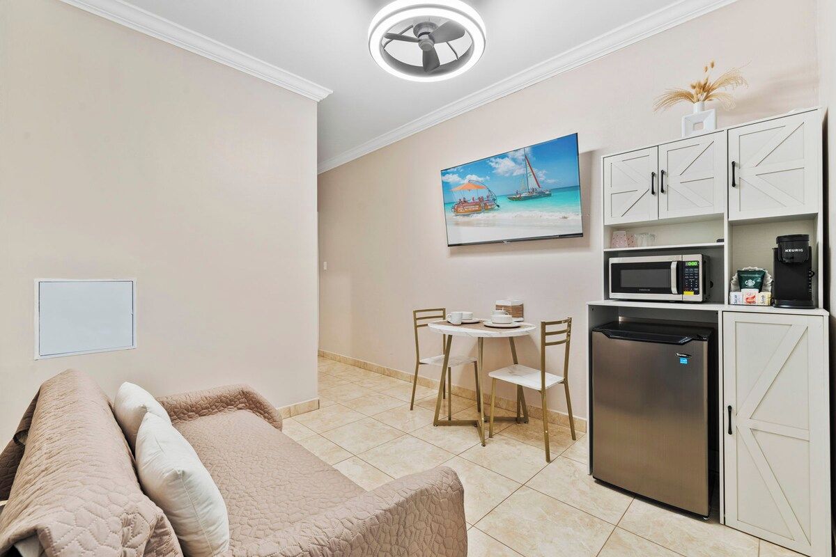 A compact room with a bed, a small dining table, chairs, a microwave, a refrigerator, and a painting of a beach scene.