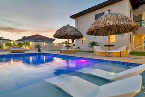 A swimming pool with lounge chairs and thatched umbrellas, surrounded by a building with a tiled roof.