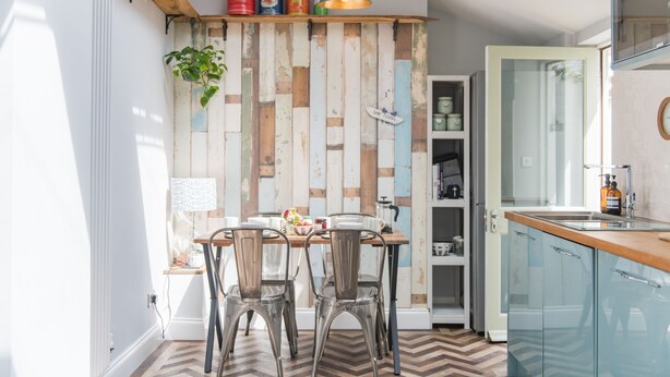 A modern kitchen with wooden panel walls, a dining area with metal chairs, and a built-in shelf with colorful containers.