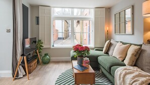 A cozy living room with a green sofa, a wooden coffee table, and a large window offering a view of a narrow street.