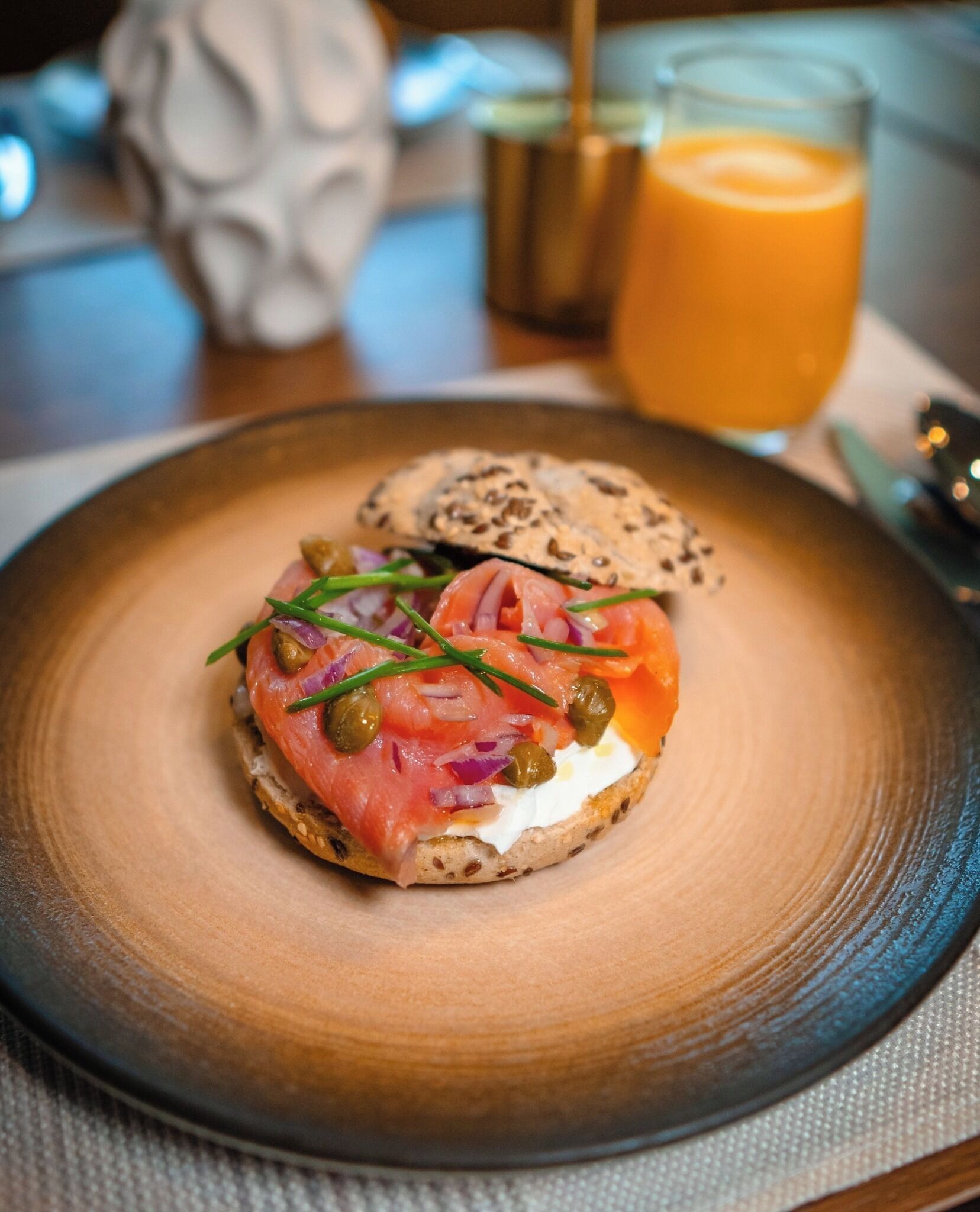 A breakfast plate with a bagel topped with smoked salmon, cream cheese, chives, and capers, accompanied by a glass of orange juice.
