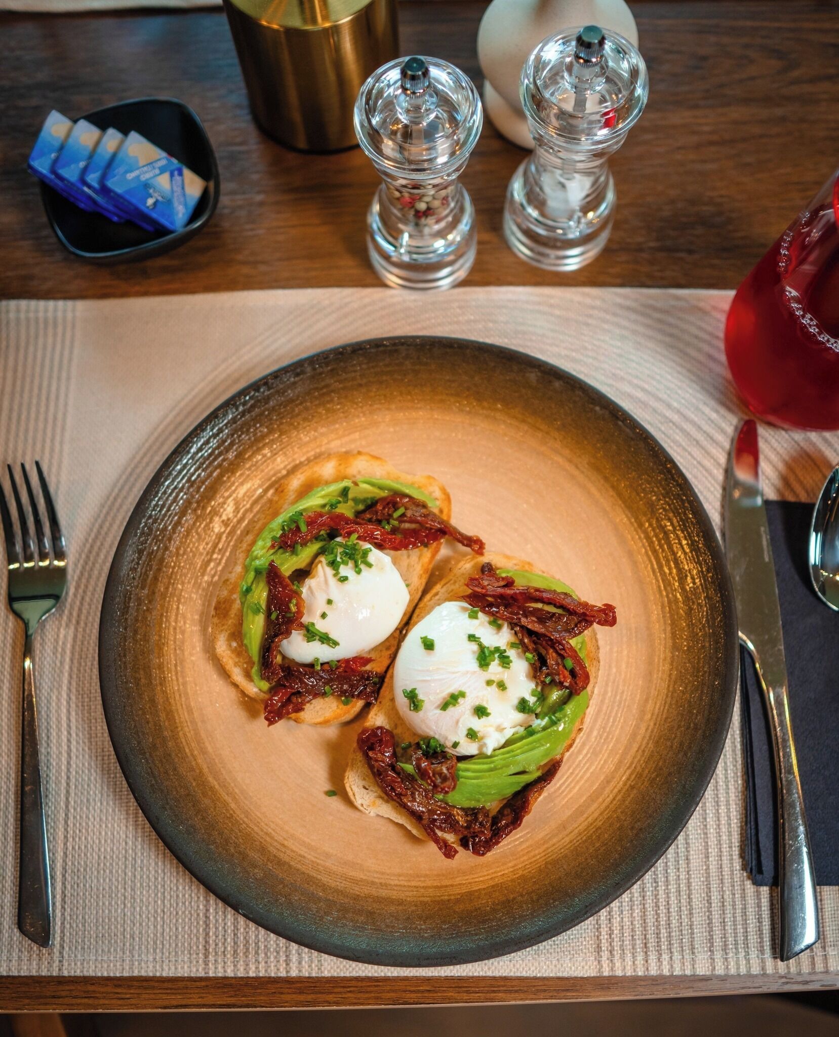 A plate with two poached eggs on toast, garnished with chives and sun-dried tomatoes, on a textured beige placemat.