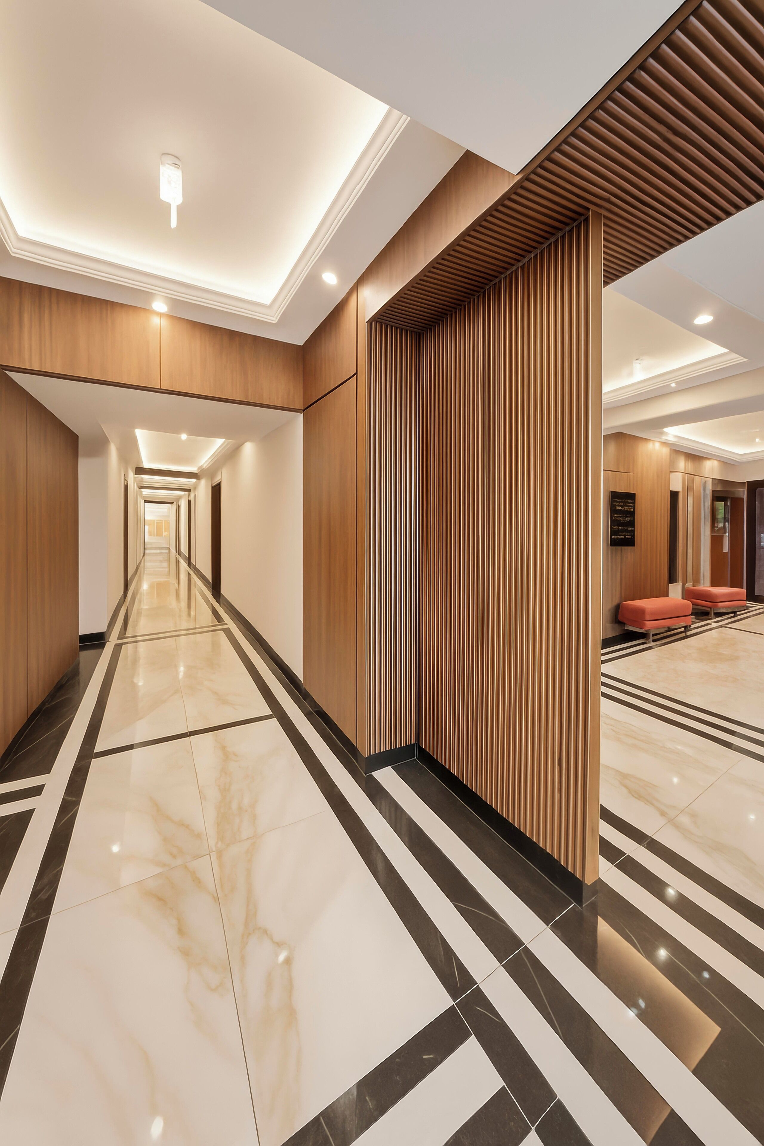 Un couloir d’hôtel moderne avec un sol en marbre poli, des panneaux en bois et un coin salon agrémenté de coussins rouges.