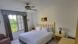 A bedroom with a bed, a chair, a nightstand, a lamp, a ceiling fan, and a window with curtains.