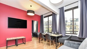 A modern living room with a red wall, a flat-screen TV, a dining table with chairs, and a grey sofa. - Authentic Apartment - 2br/6p - Near Opera Garnier (Paris)