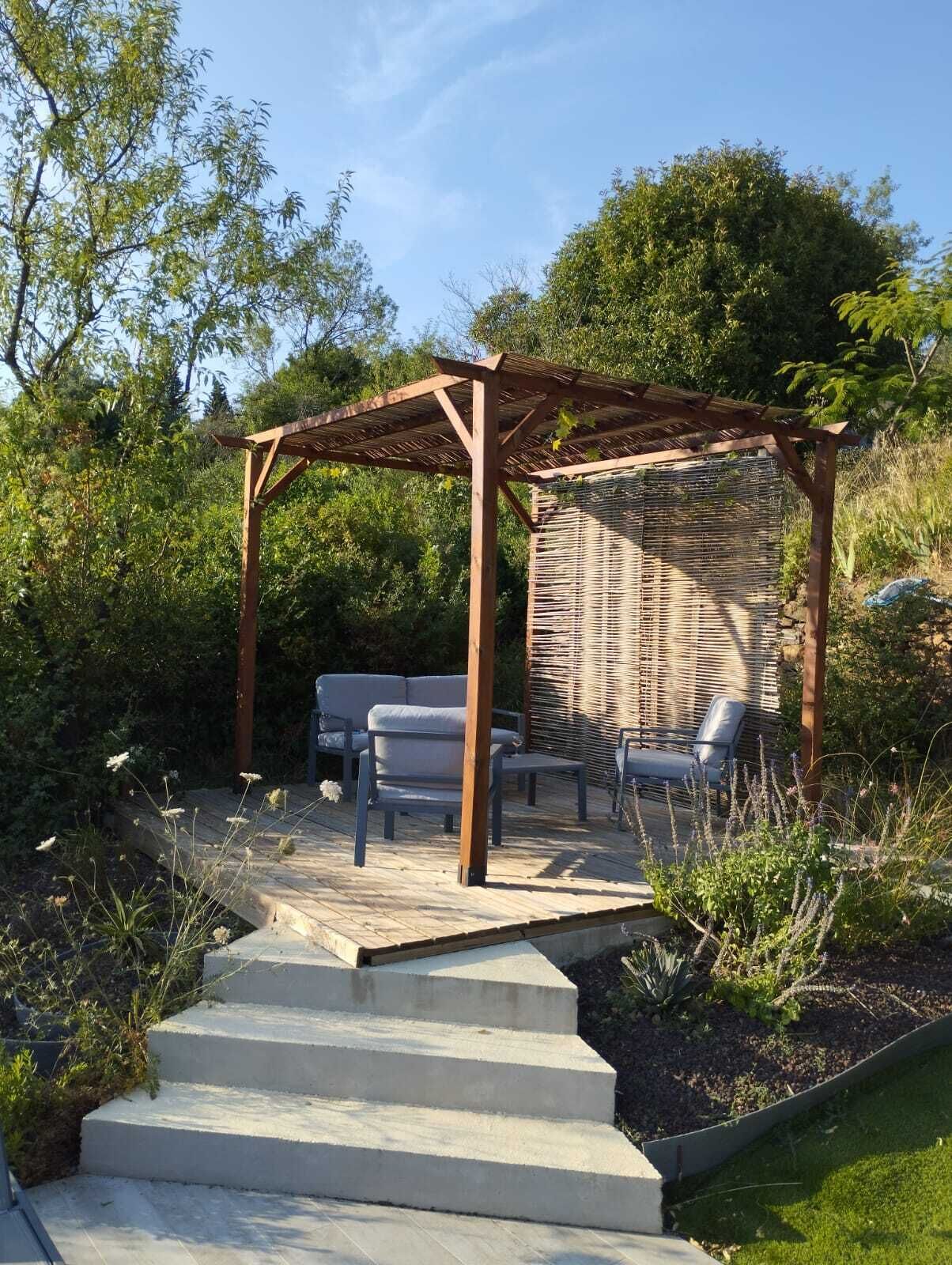 Eine Pergola aus Holz mit einer gewobenen Wand, Sitzgelegenheiten im Freien und einer Betontreppe, die hinauf führt.