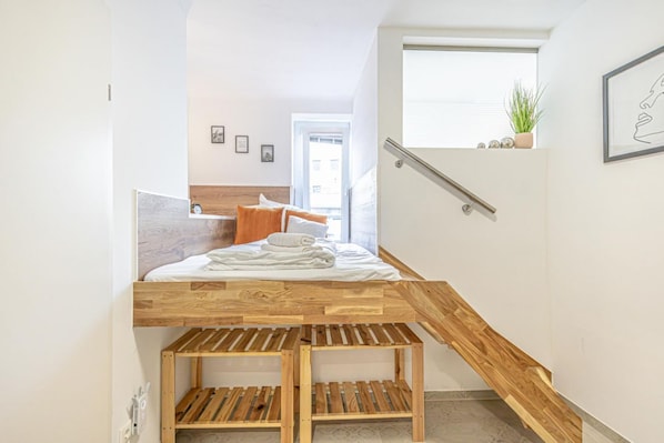 A bedroom with a wooden bunk bed, a bedside table, a window with blinds, and a small plant on the windowsill.