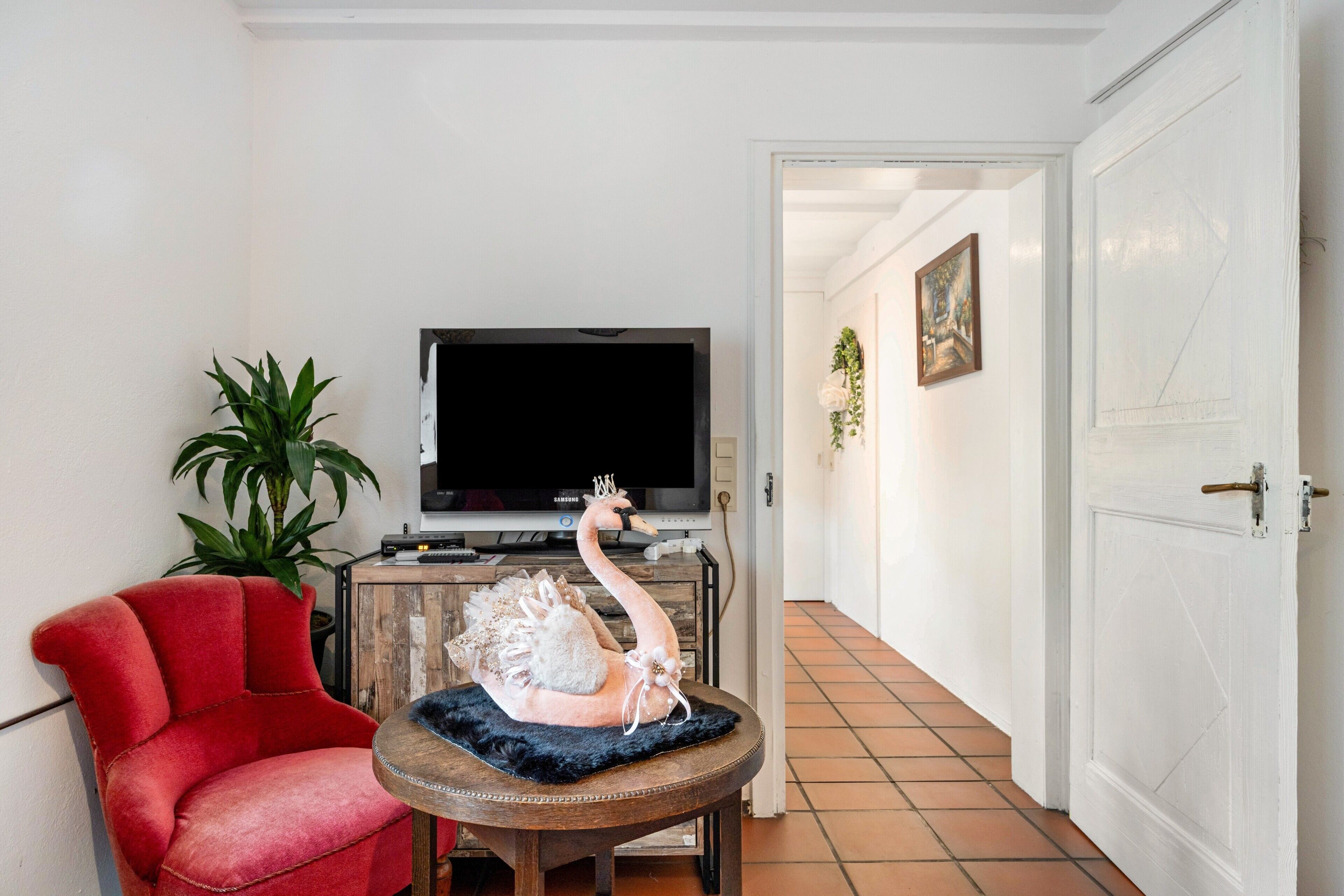 A living room with a red chair, a round table, a decorative swan, a television, and a door leading to another room.