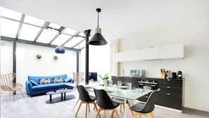 A modern living room with a glass dining table, black chairs, a blue sofa, and a kitchen area with white cabinets and a black countertop. - Elegant Apartment -2br/4p - Sacre-coeur (Paris)