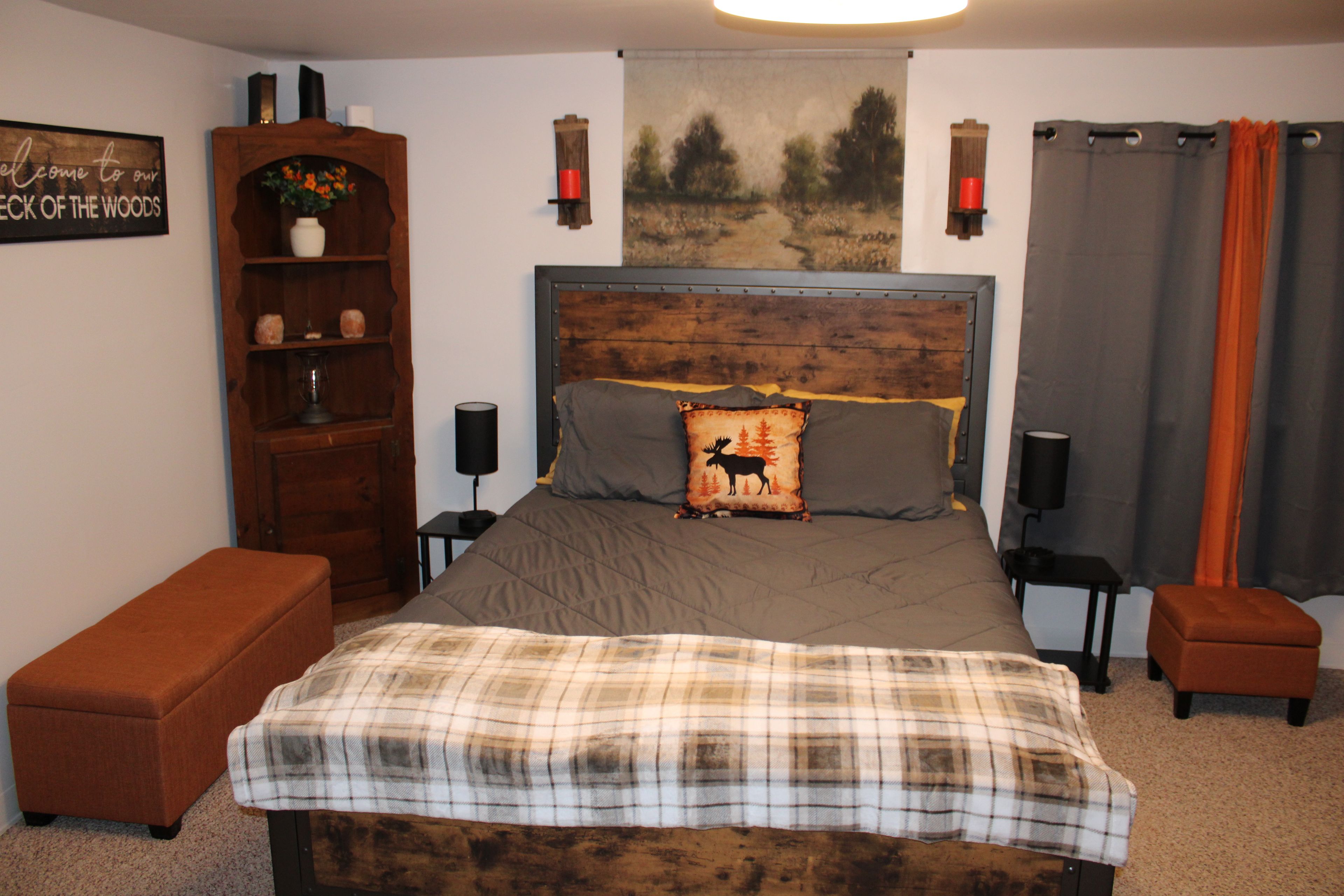 Un dormitorio con cabecera de madera, cama con colcha a cuadros, mesita de noche con lámpara, un banco, estantería y un cartel en la pared.