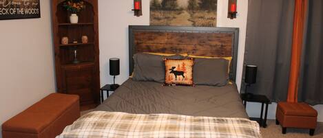 Un dormitorio con cabecera de madera, cama con colcha a cuadros, mesita de noche con lámpara, un banco, estantería y un cartel en la pared.