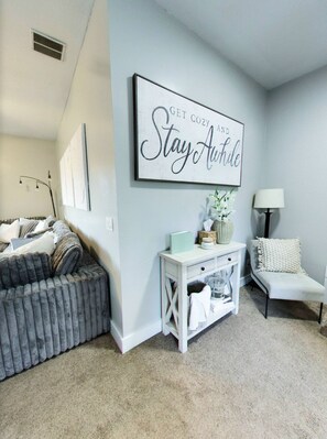 A cozy room with a gray sofa, a white side table, and a chair. A large sign on the wall reads "GET COZY AND Stay Awhile." - Vernal Sport Court Base Camp (Vernal)