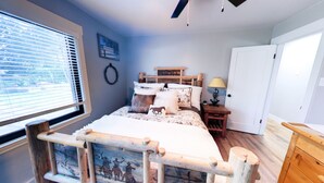 A bedroom with a bed, a bedside table, a lamp, a window with blinds, and a door.