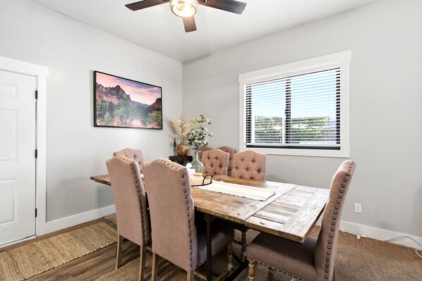 A dining room with a wooden table, six upholstered chairs, a ceiling fan, a window with blinds, a painting on the wall, and a door. - Vernal Sport Court Base Camp (Vernal)
