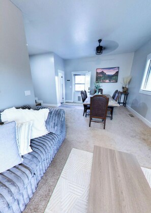 A spacious living room with a grey sofa, a dining area with a wooden table and chairs, and a hallway leading to a door.