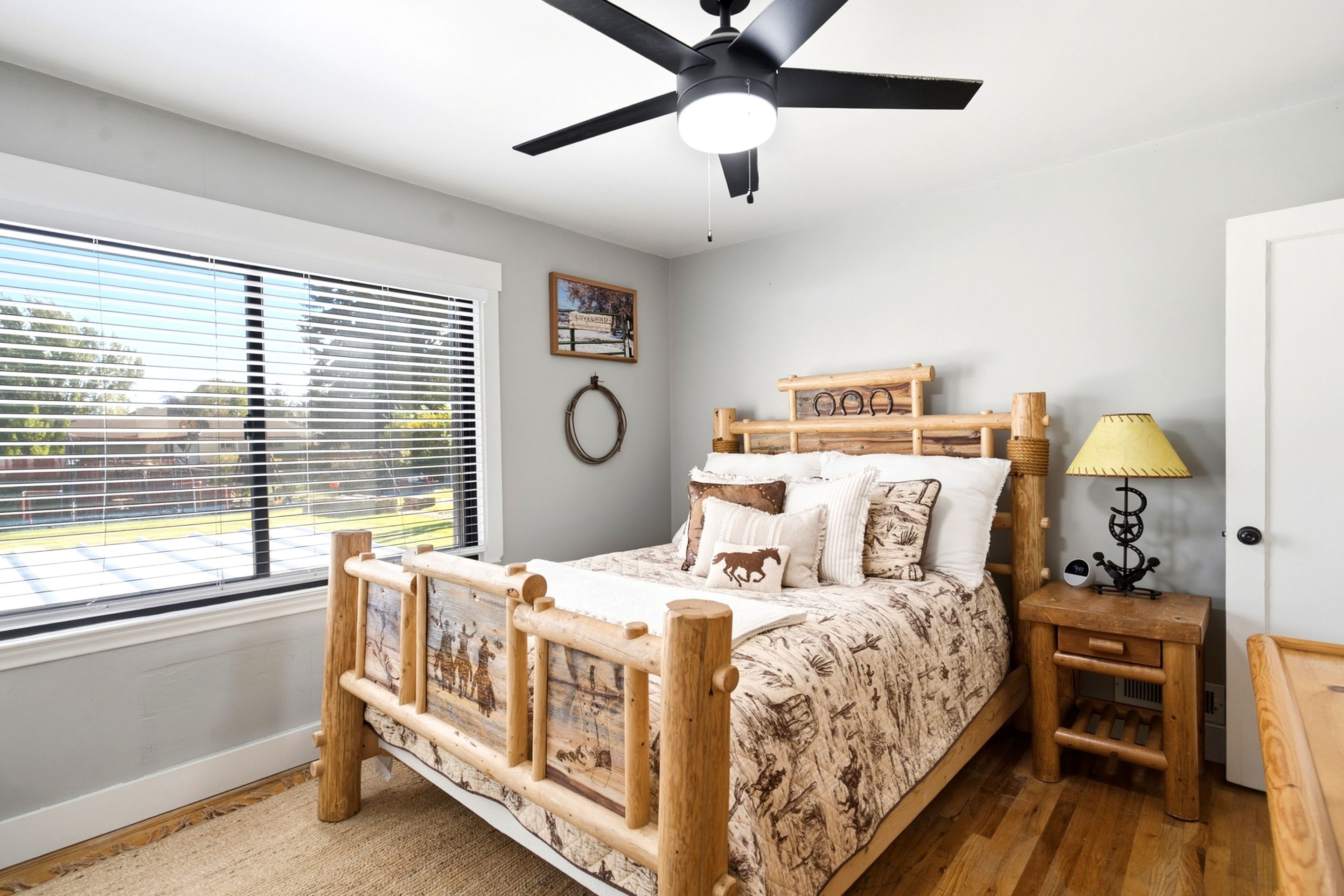 A bedroom with a bed, a bedside table, a lamp, a ceiling fan, a window with blinds, and a wall decoration.