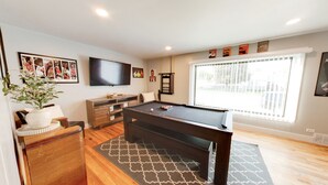 A modern living room with a pool table, a flat-screen TV, and a large window with blinds. - Vernal Sport Court Base Camp (Vernal)